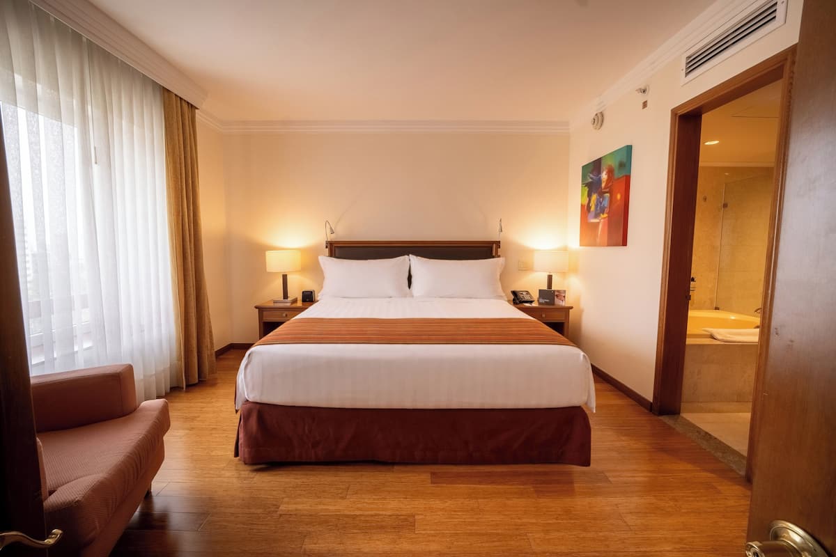 A hotel room with a large bed, two bedside lamps, a wooden floor, a chair, and a painting on the wall.