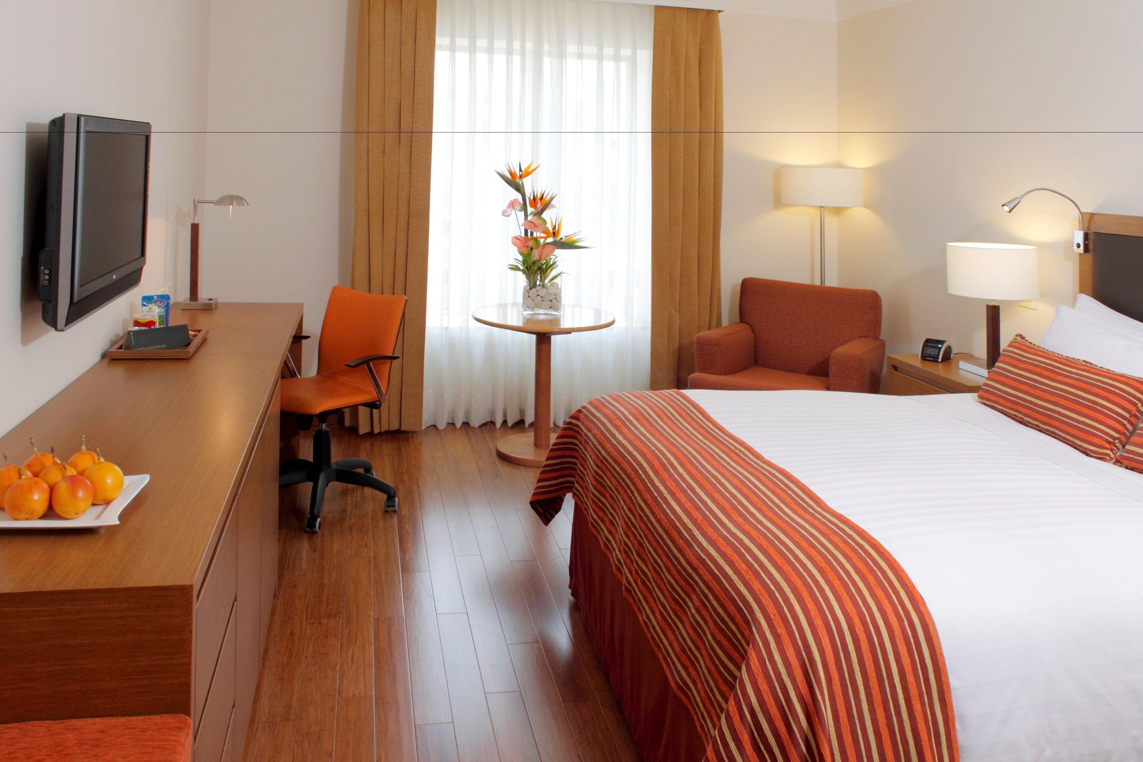 A hotel room with a bed, a desk with a chair, a TV, a lamp, and a small table with a vase of flowers.