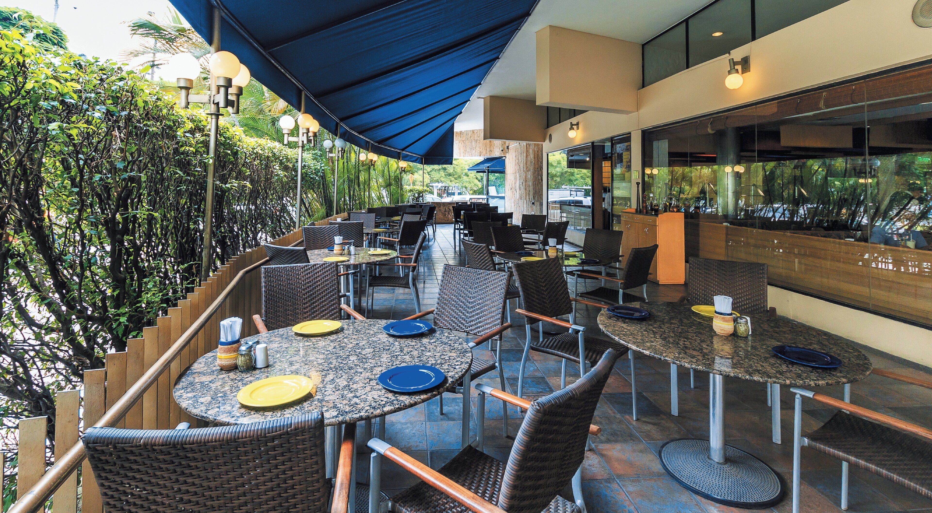An outdoor dining area with round tables and wicker chairs, surrounded by greenery and covered by blue canopies.