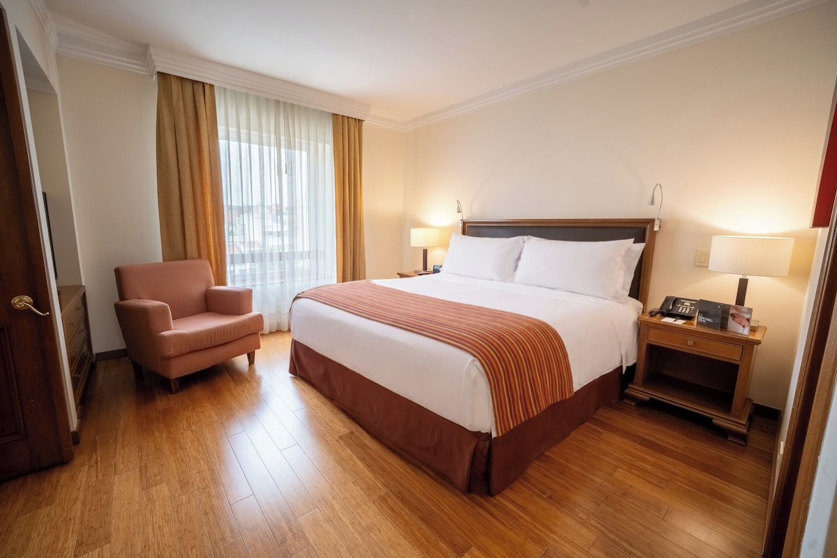 A hotel room with a bed, a chair, two bedside tables with lamps, and a window with curtains.