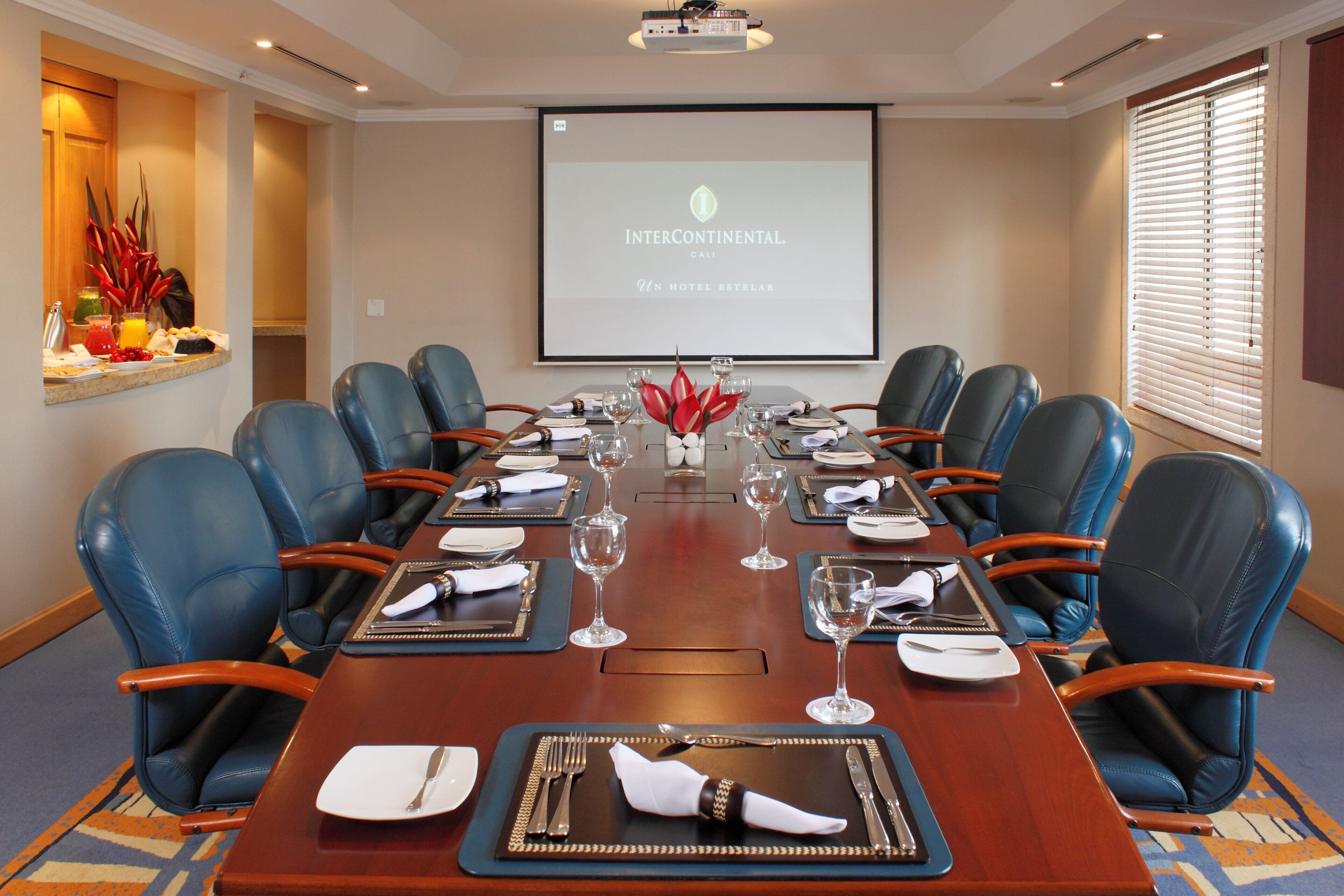 A conference room with a long table set for a meeting, blue leather chairs, a projector screen displaying 'INTERCONTINENTAL. CALI,' and a buffet area with fruits and beverages.