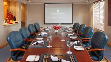 A conference room with a long table set for a meeting, blue leather chairs, a projector screen displaying 'INTERCONTINENTAL. CALI,' and a buffet area with fruits and beverages.