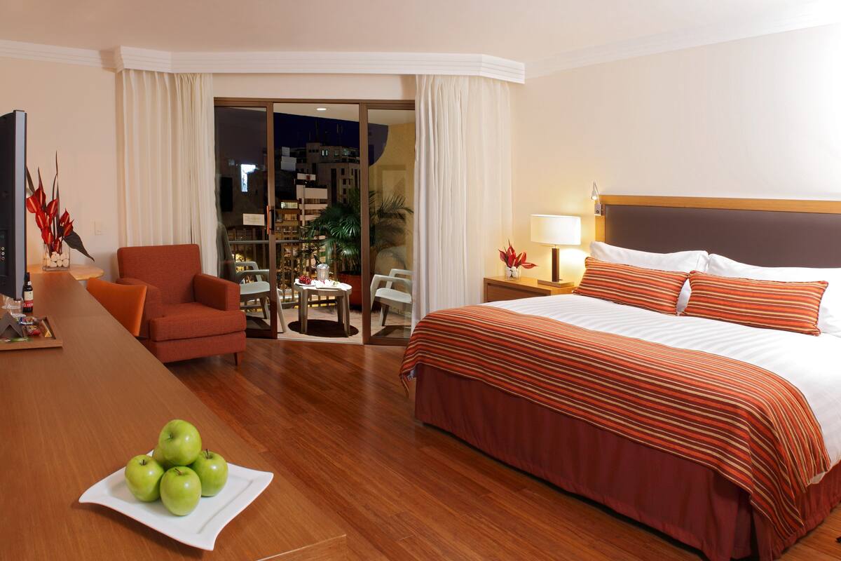 A hotel room with a bed, a desk with apples, a chair, and a view of the outside.