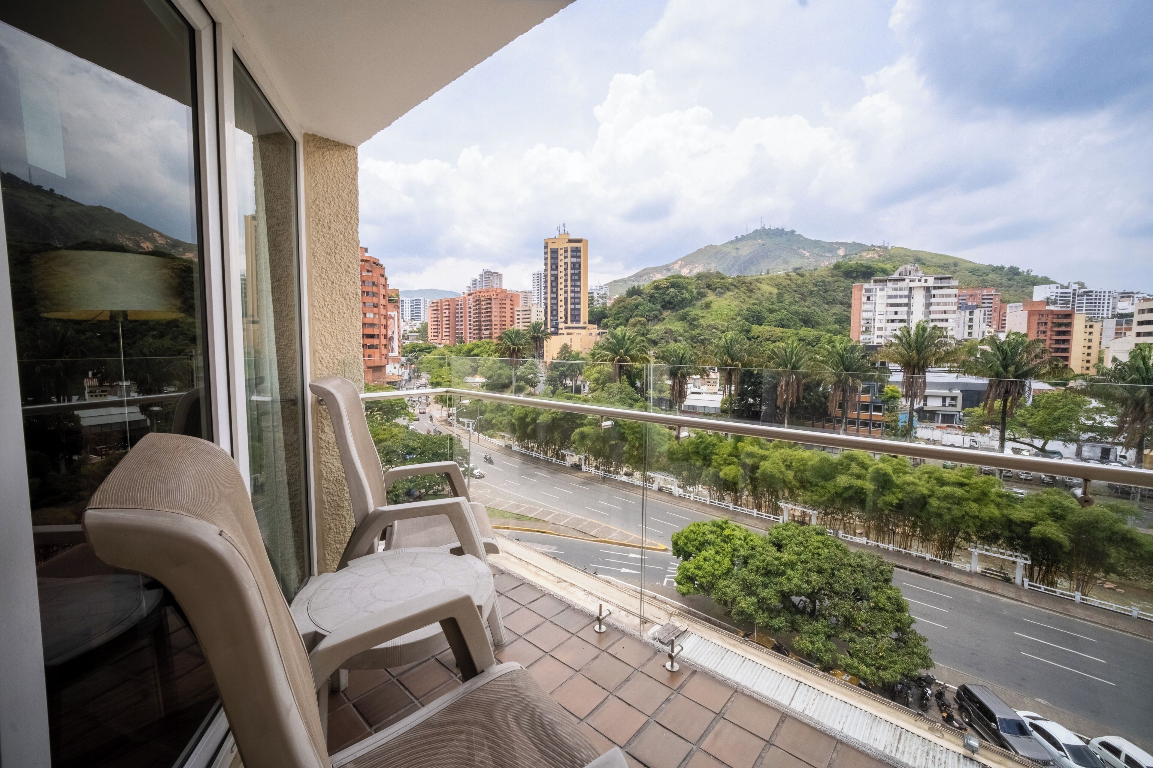 A balcony with chairs, a view of a cityscape with buildings and a highway, and a hilly background.