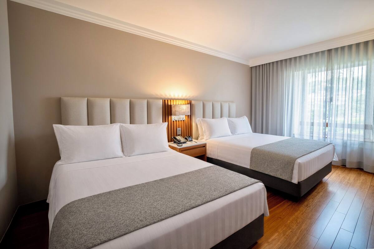 A hotel room with two beds, a bedside table with a lamp, and a large window with curtains.