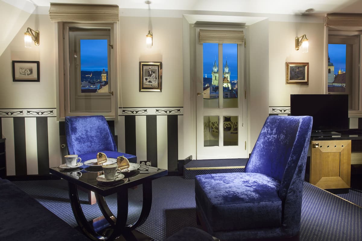A hotel room with a view of a city skyline, a dining table with a cake, two blue armchairs, and a television.