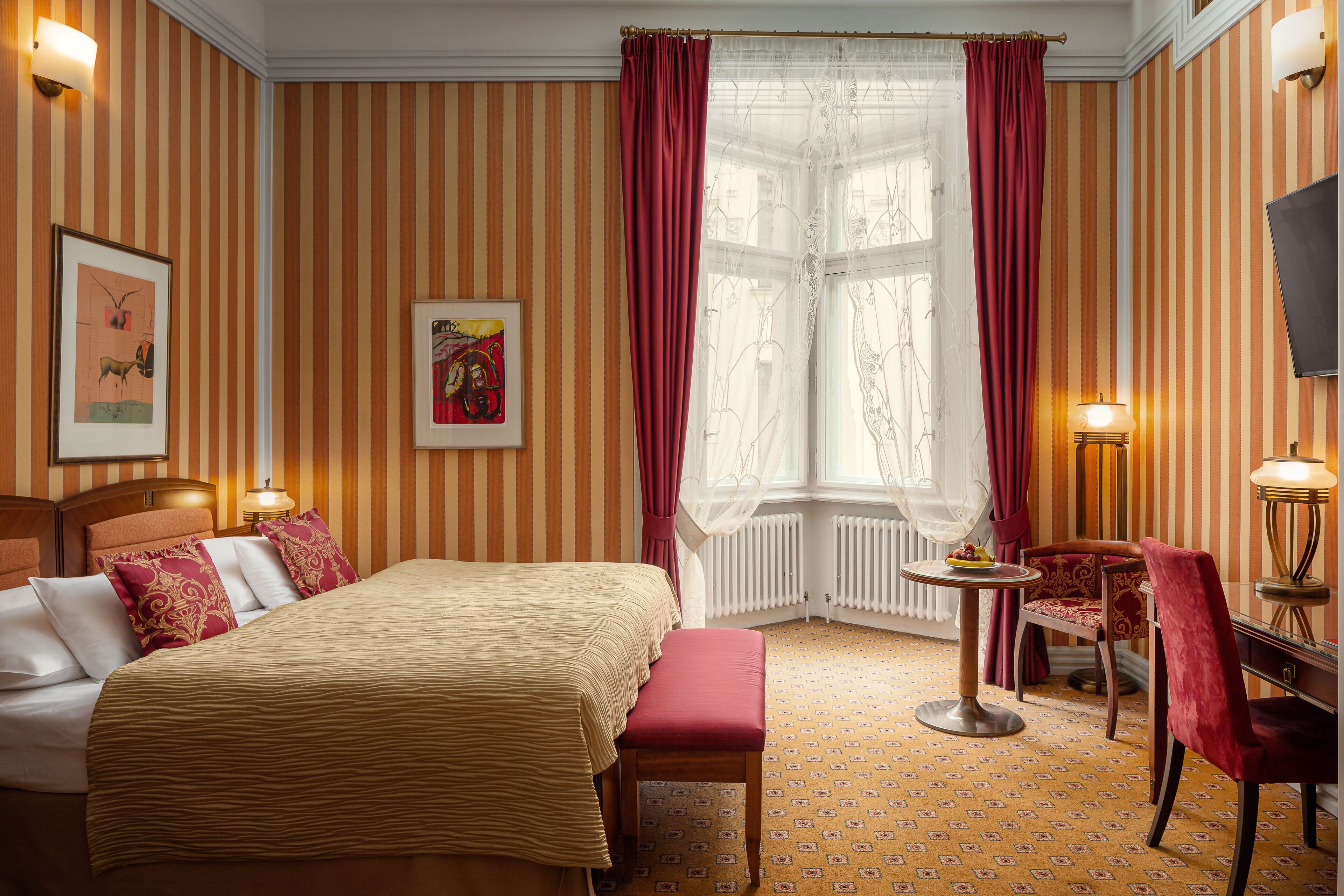 A hotel room with a bed, a bench, a small table, a chair, and a window with curtains.