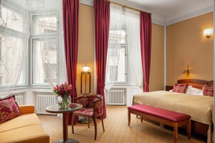 Quarto de hotel com cama, um banco, mesa com um vaso de flores, uma cadeira e janela com cortinas.