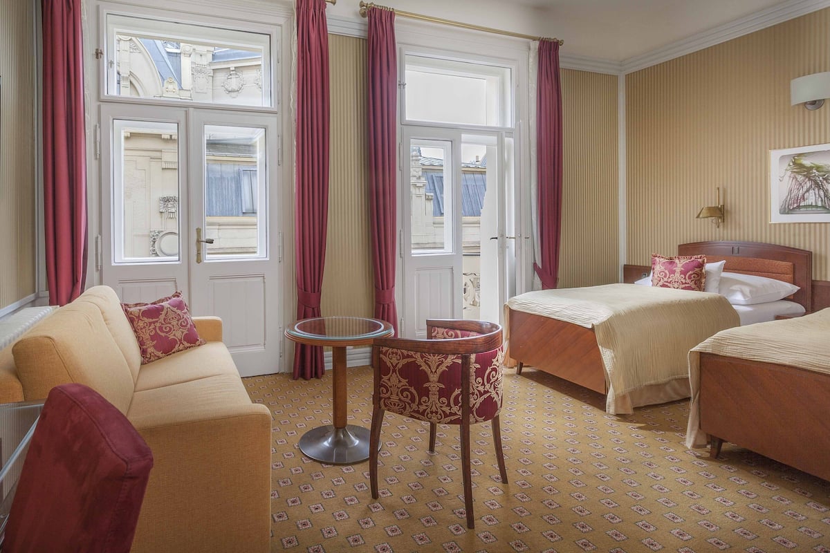 A hotel room with a sofa, two beds, a small table, and a view through the windows.