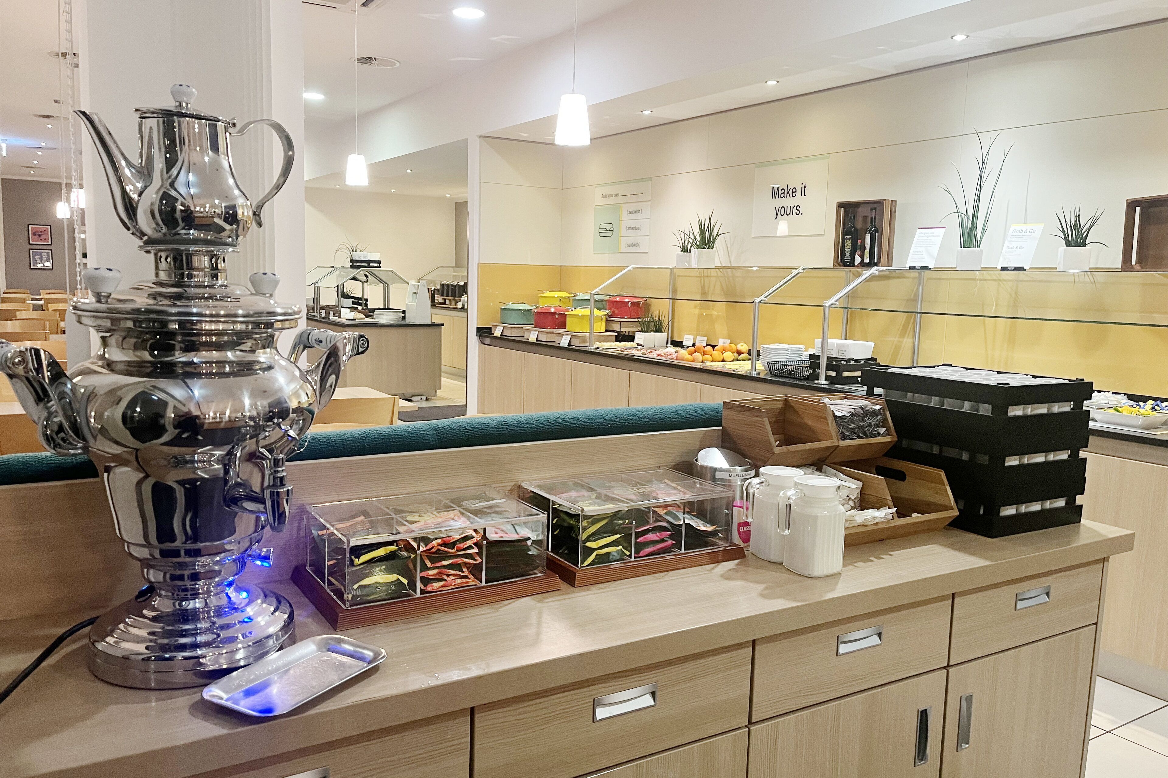 A modern hotel reception area with a polished metal coffee urn, clear plastic storage bins, and a variety of amenities including a coffee machine, a scale, and a display of bottled water.
