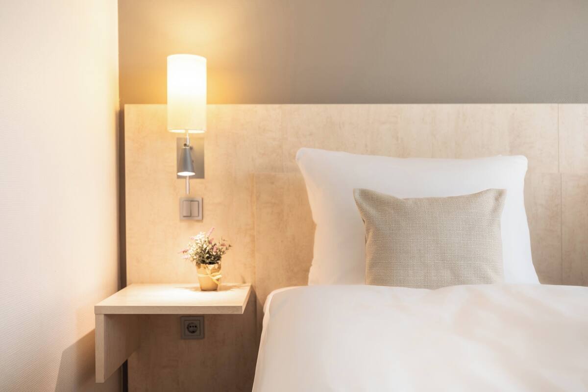 A neatly made bed with white linens and a single decorative pillow, a bedside table with a lamp and a small vase of flowers, and a wall-mounted light fixture.