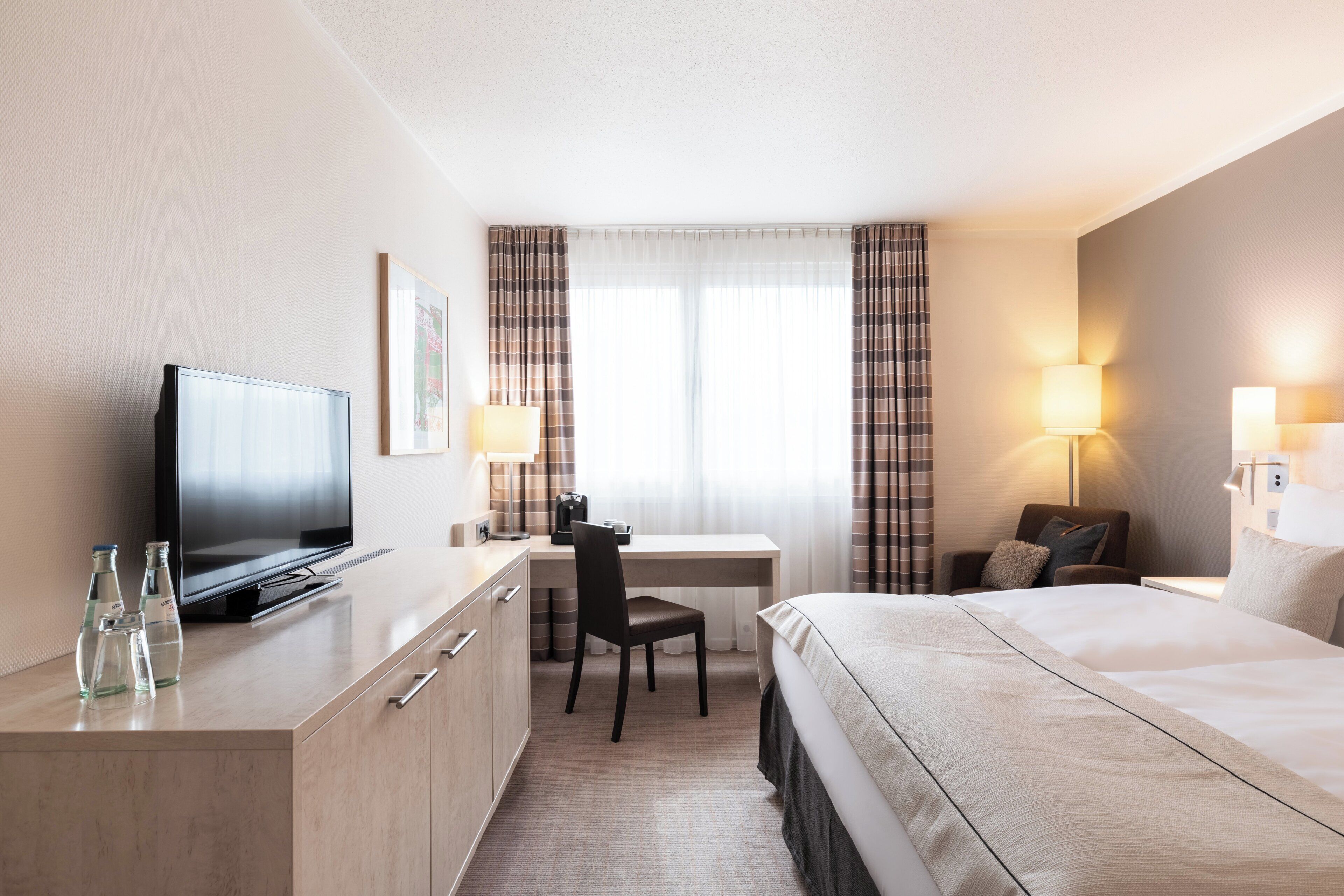 A hotel room with a bed, a desk with a chair, a television, and a window with curtains.