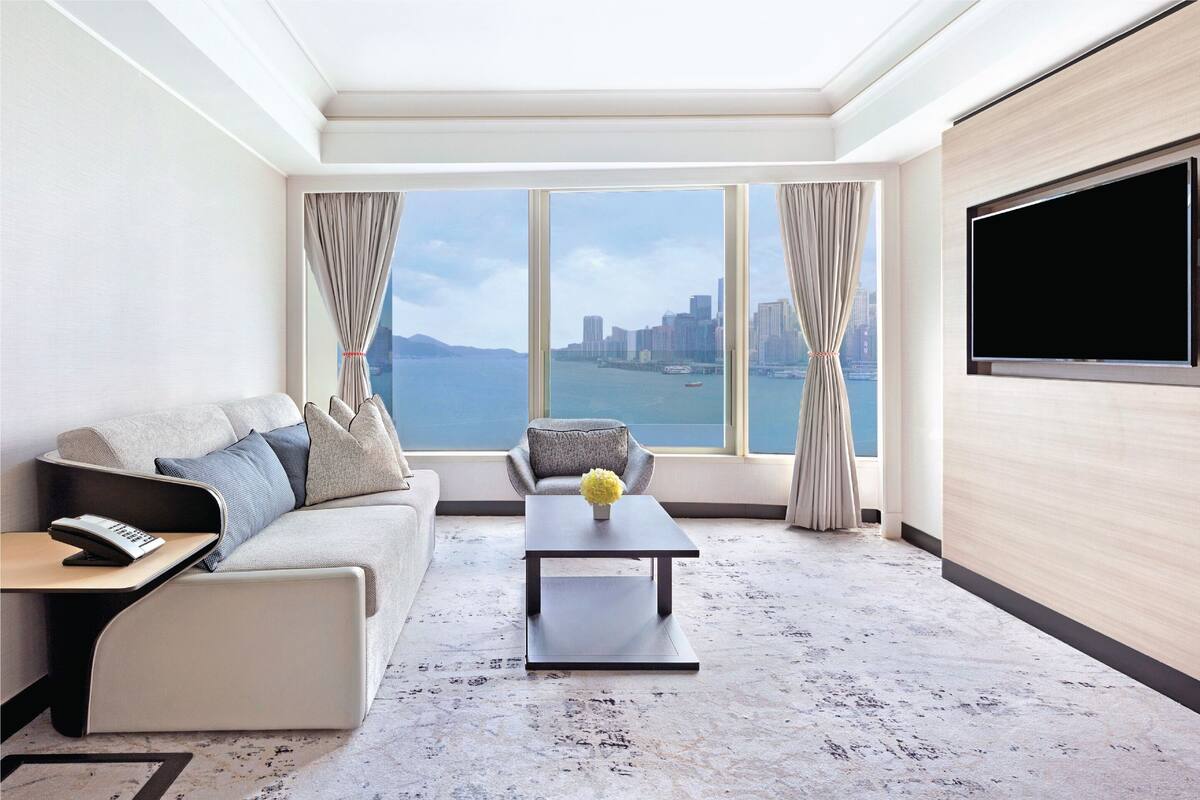 A modern hotel room with a sofa, a coffee table, a TV, and a view of a cityscape through large windows.