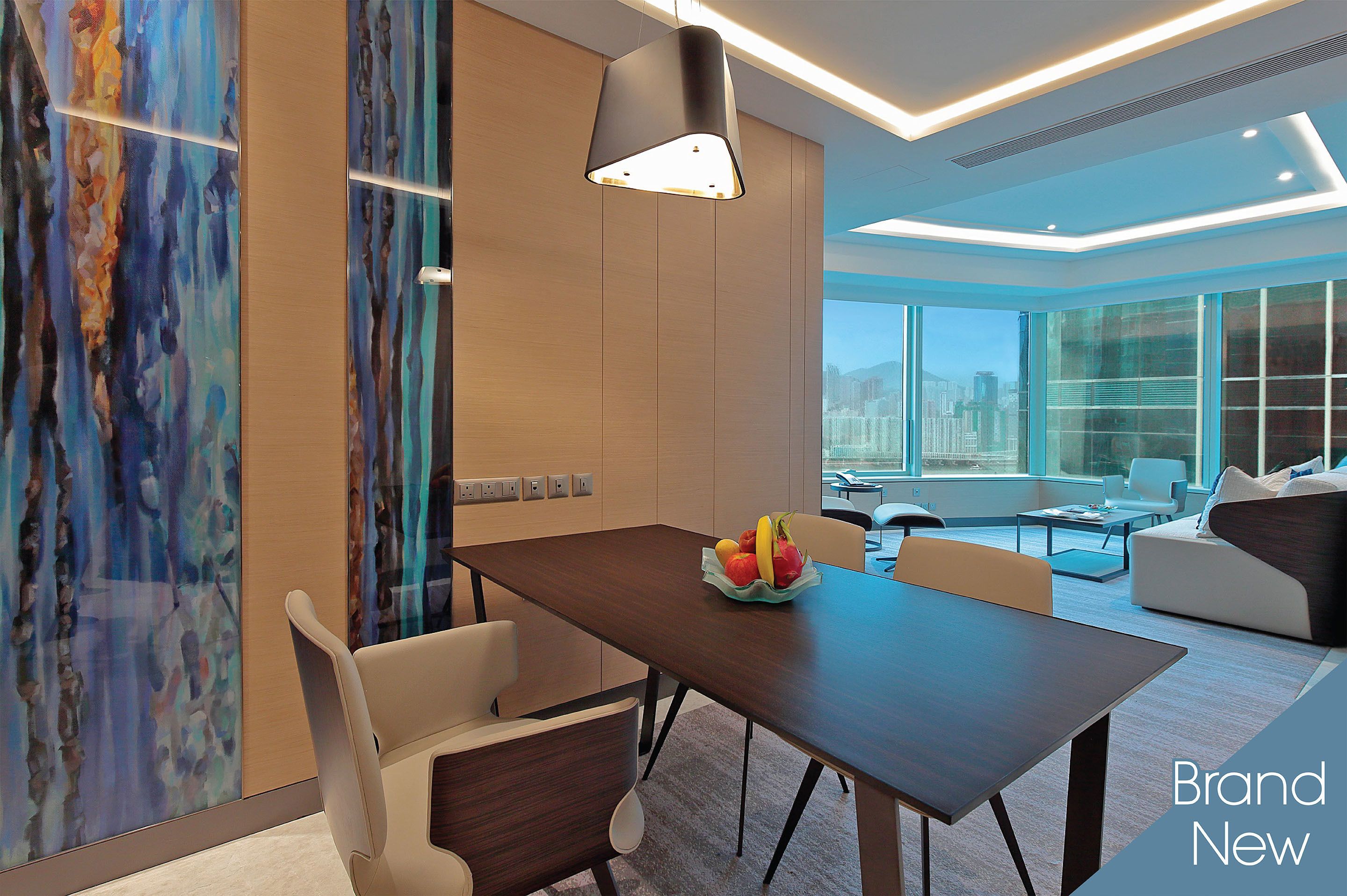 A modern dining area with a wooden table, white chairs, and a fruit bowl. There is a large window with a city view, and the room has a contemporary design with a ceiling light fixture.