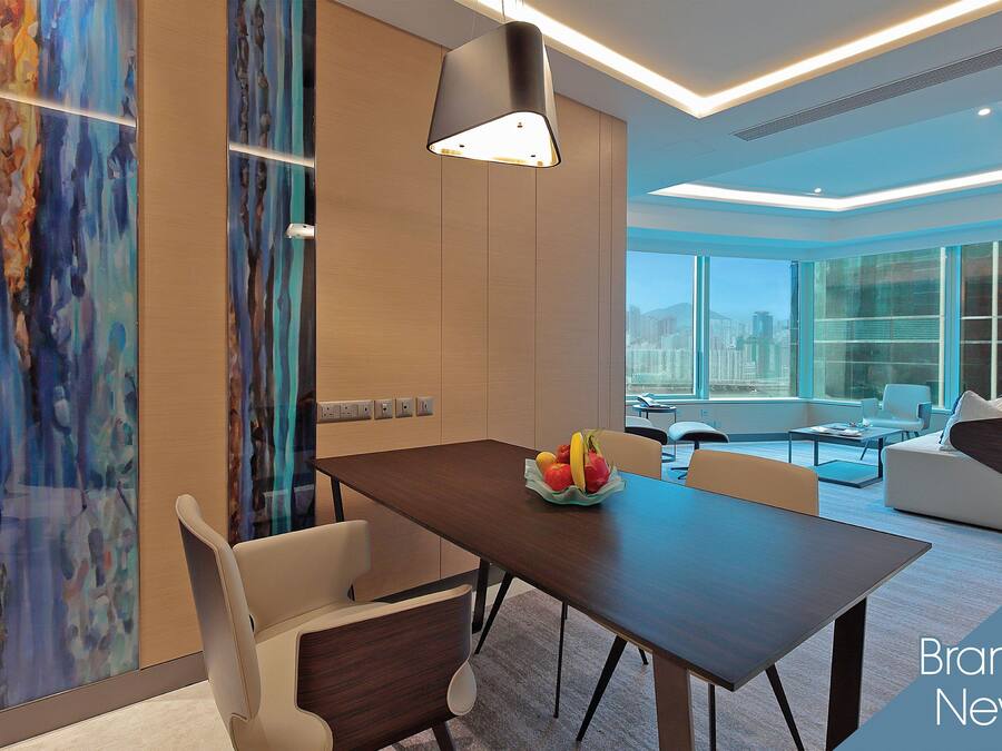 A modern dining area with a wooden table, white chairs, and a fruit bowl. There is a large window with a city view, and the room has a contemporary design with a ceiling light fixture.