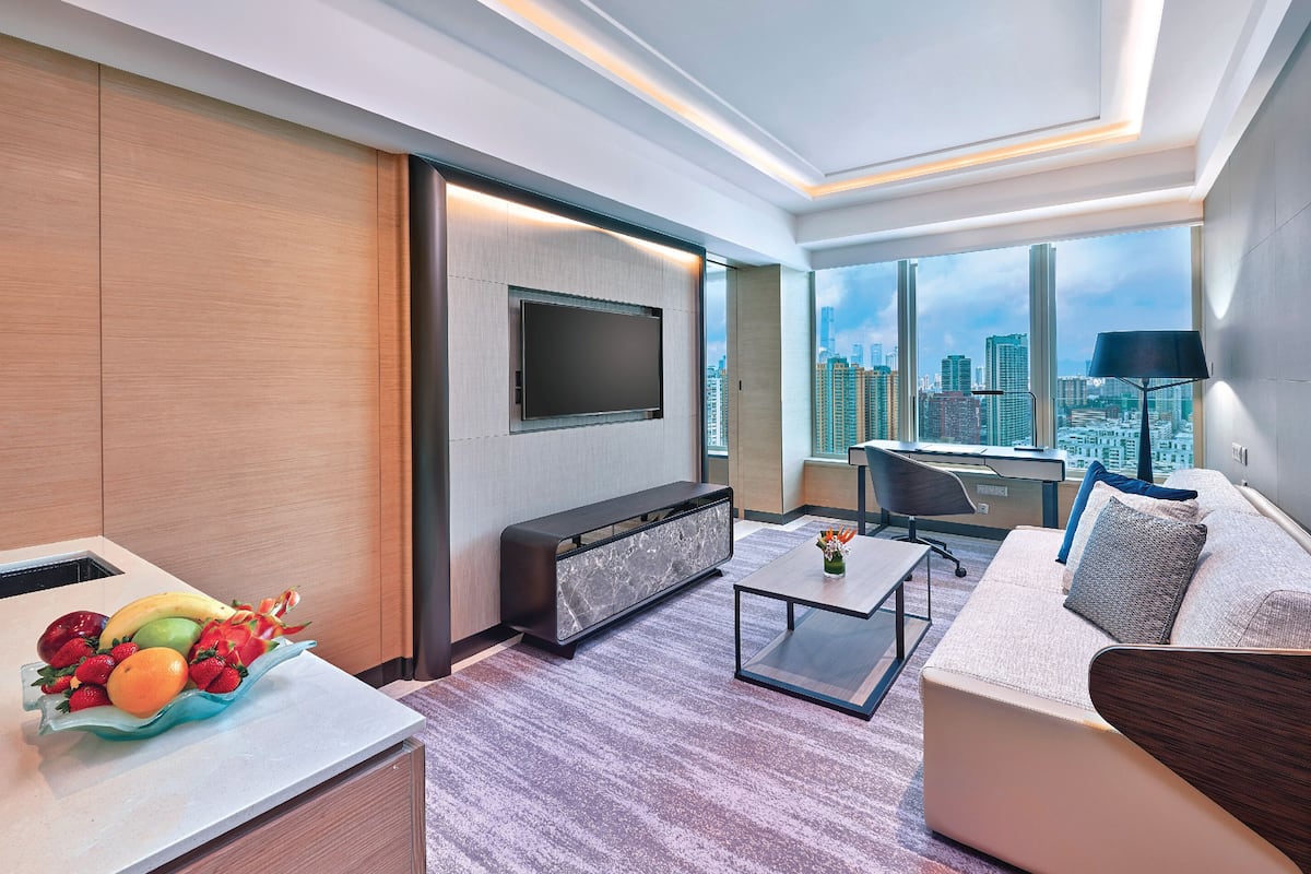 A modern hotel room with a large window, a flat-screen TV, a sofa, a coffee table, and a fruit bowl.
