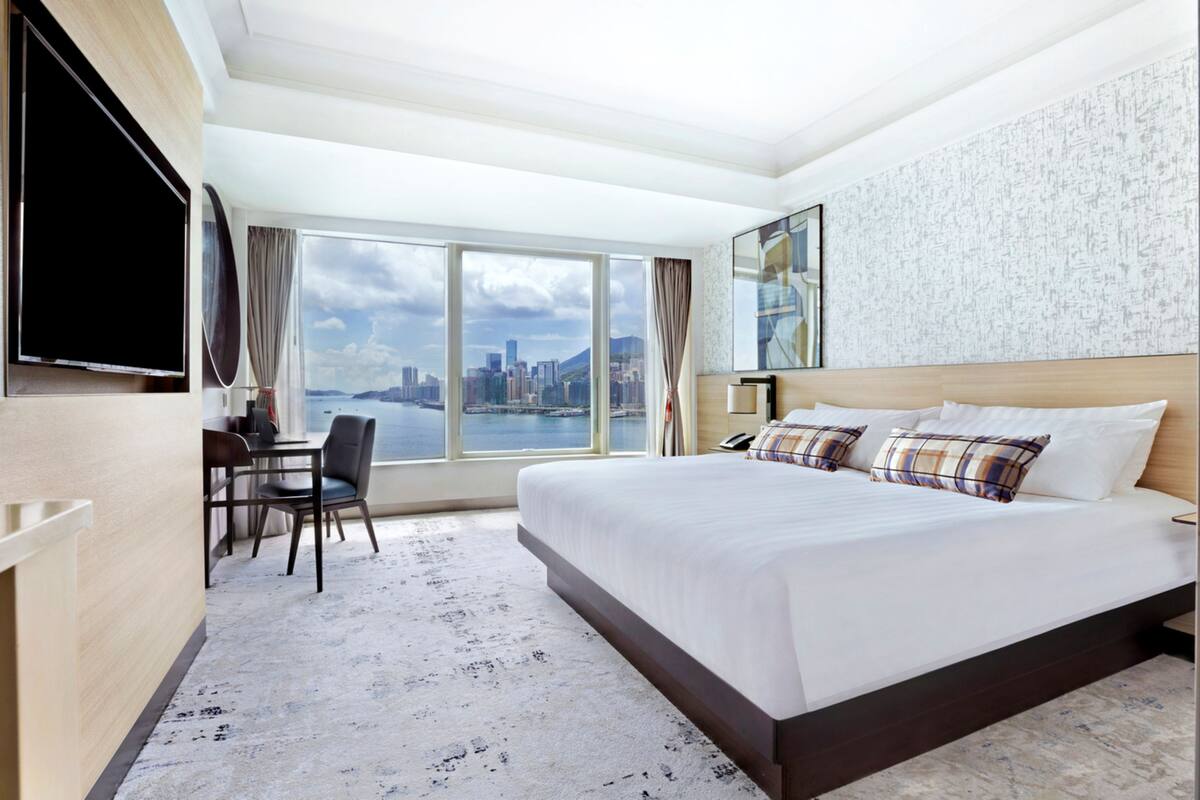 A hotel room with a large bed, a desk with a chair, a TV mounted on the wall, and a large window offering a city view.