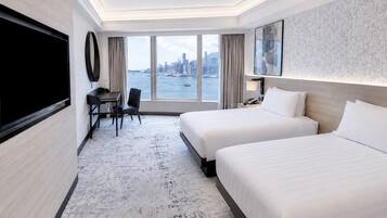 A hotel room with two beds, a desk, a chair, a TV, and a large window with a city view.