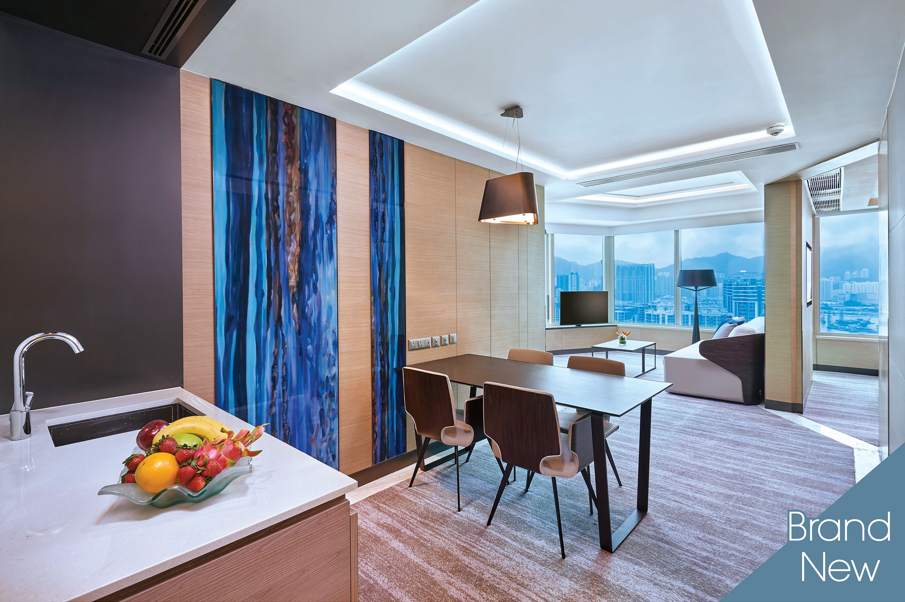 A modern hotel room with a dining area, a fruit bowl on the counter, and a city view.