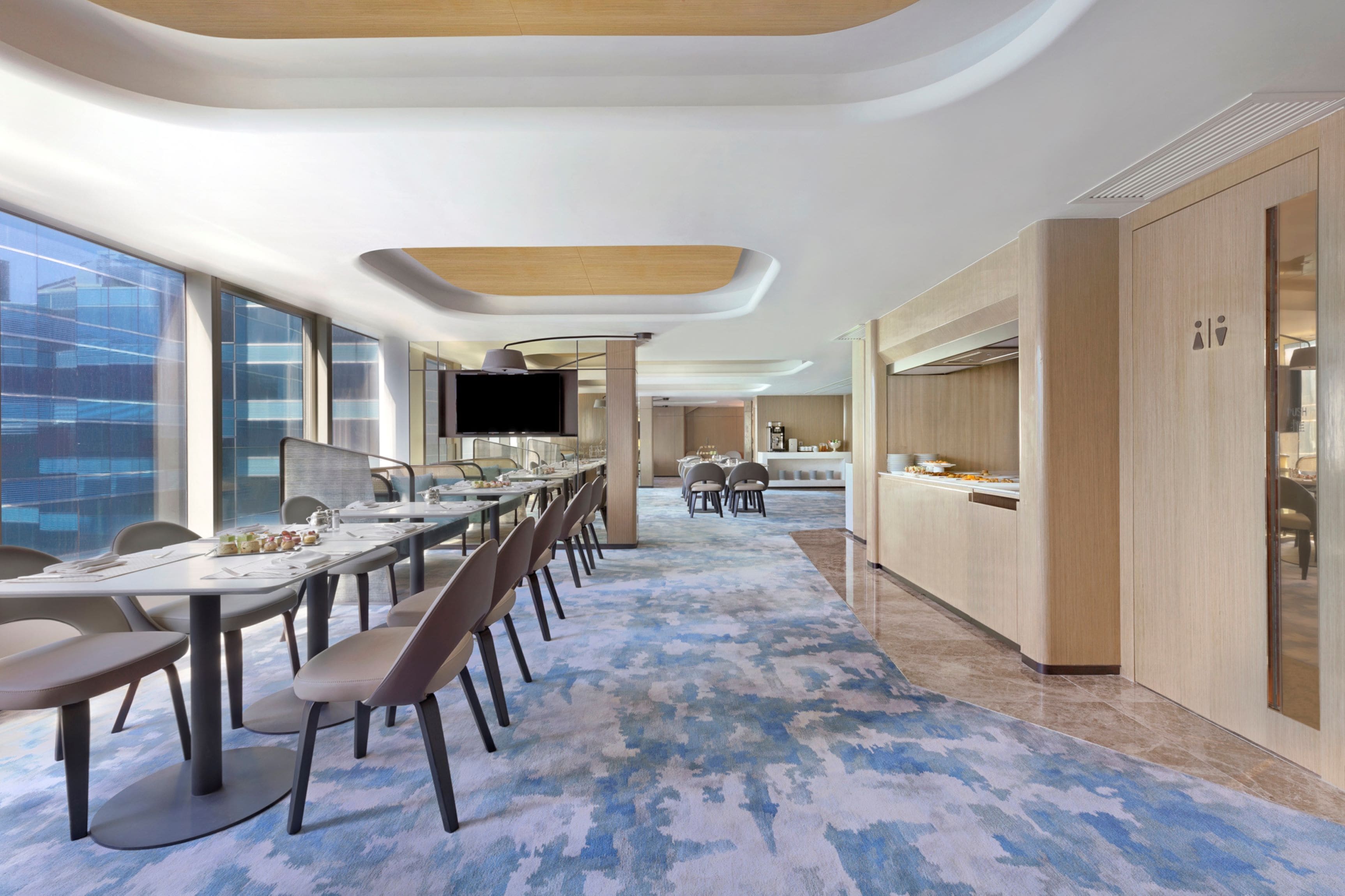 A modern hotel dining area with long tables, chairs, and a large TV screen.