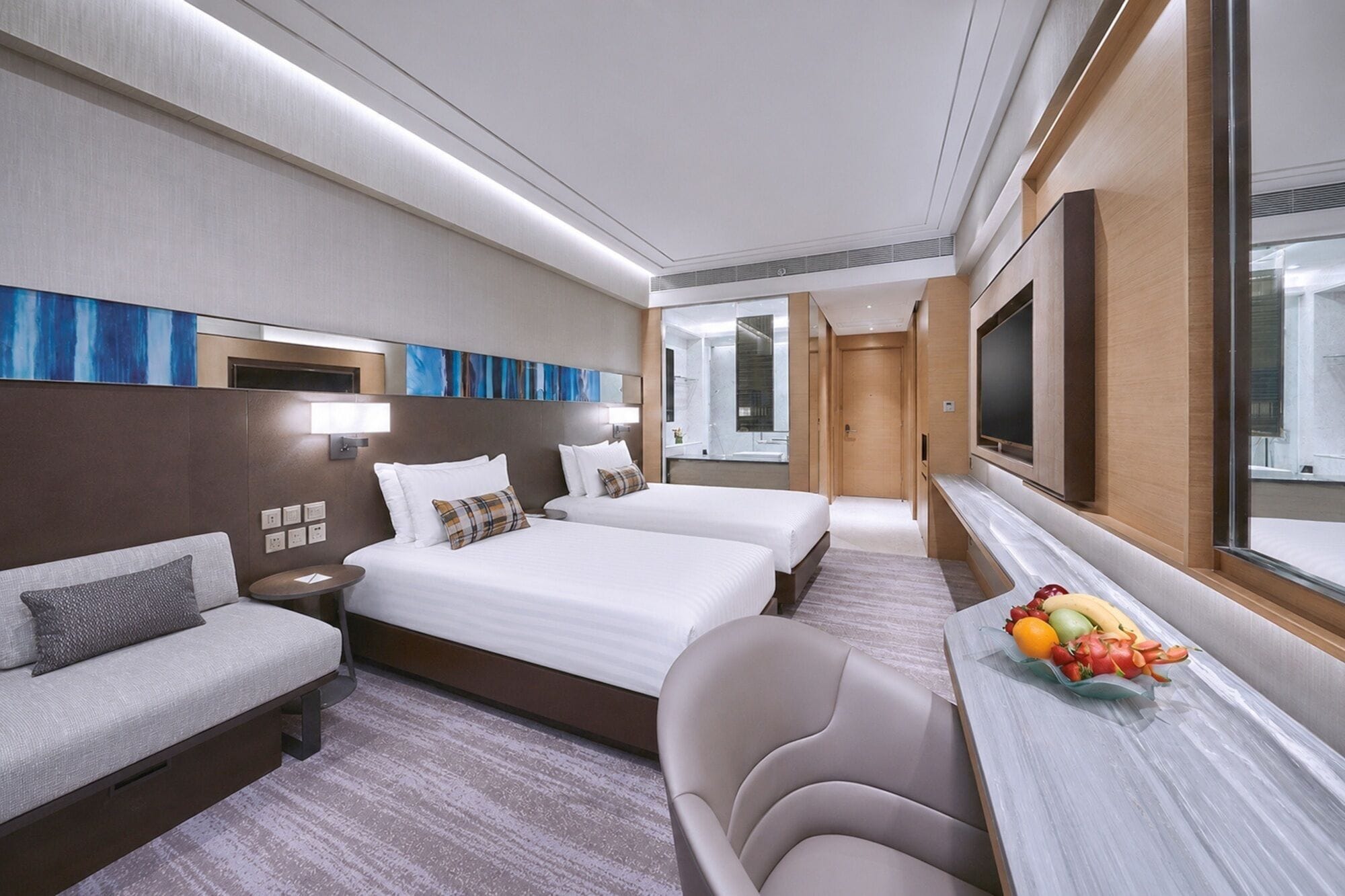 A hotel room with two beds, a sofa, a desk with a fruit bowl, and a TV.