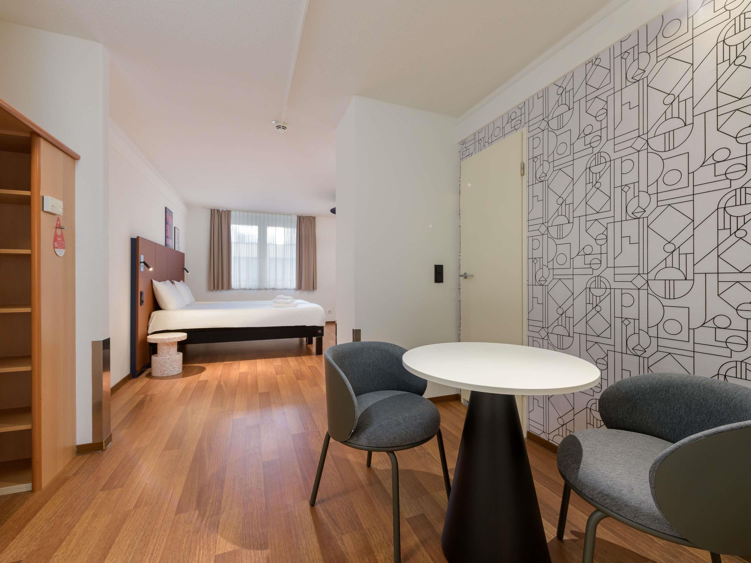 A hotel room with a bed, two chairs, a small table, and a wardrobe.
