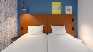Et hotelværelse med et moderne design, der indeholder en stor seng med hvide sengetøj, en sengegavl med geometrisk mønster og en væg med abstrakt kunst.