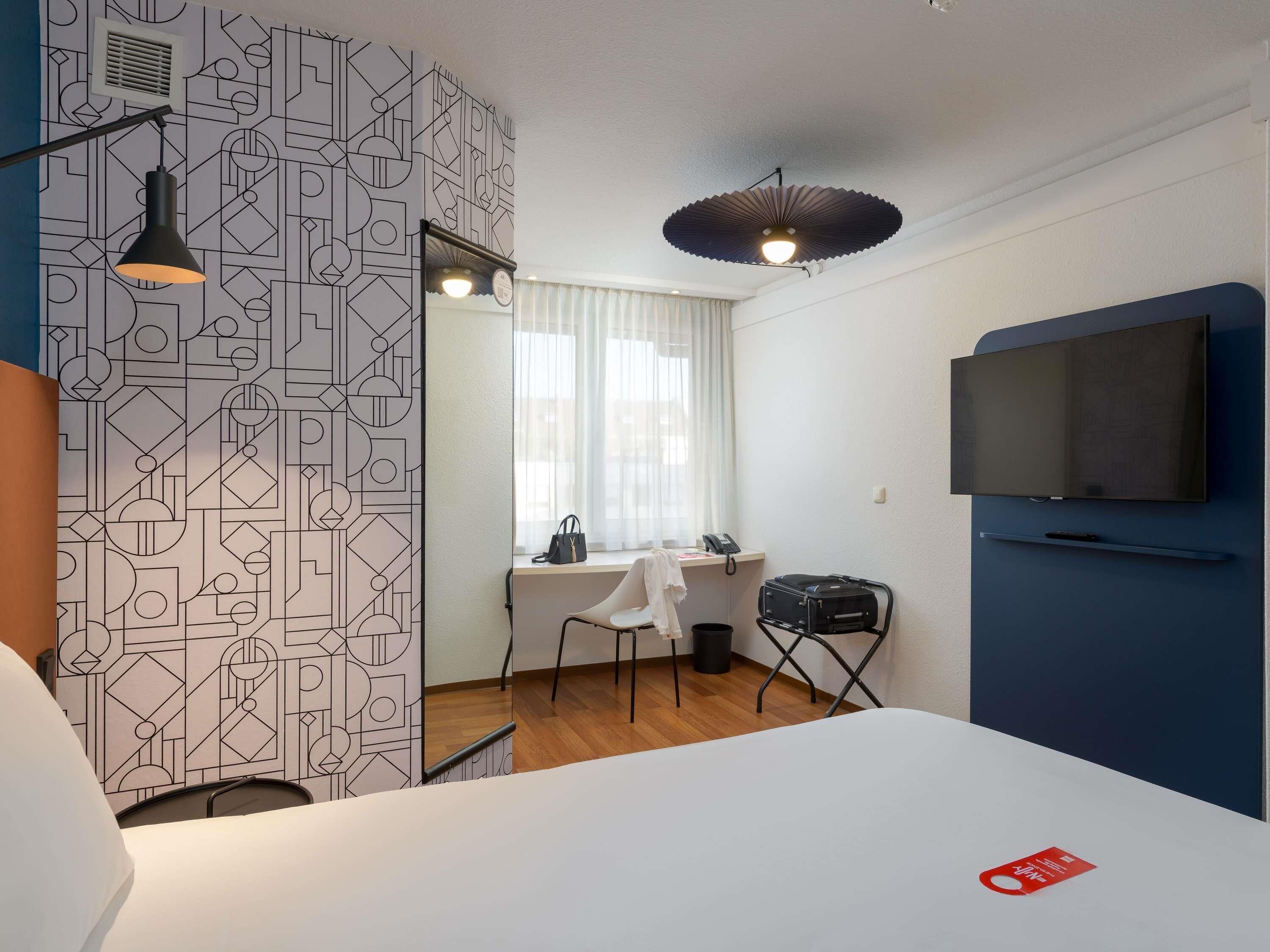 Habitación de hotel con cama, escritorio y silla, televisor montado en la pared y papel tapiz estampado.