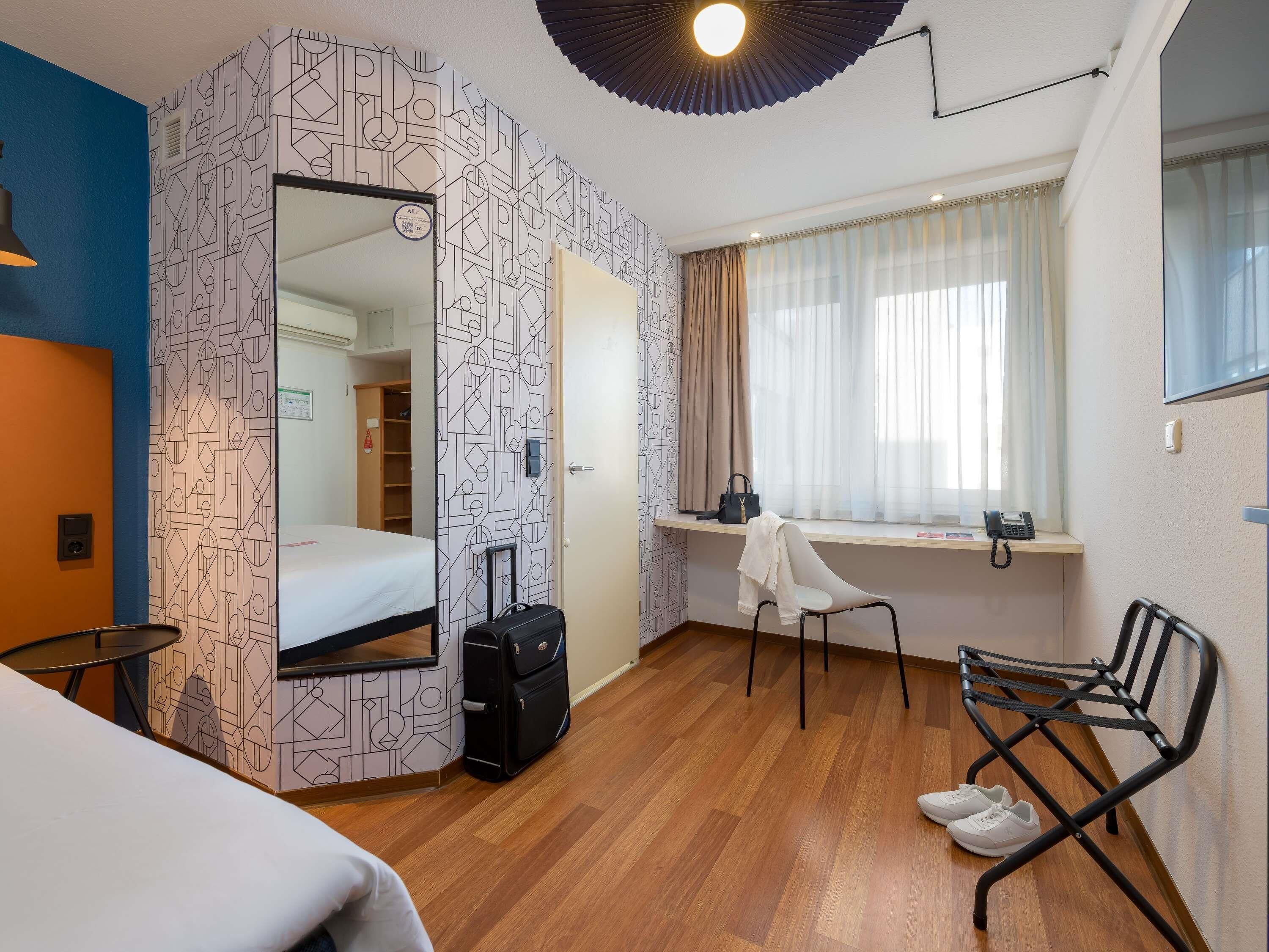 A hotel room with a bed, a desk with a chair, a suitcase, and a window with curtains.