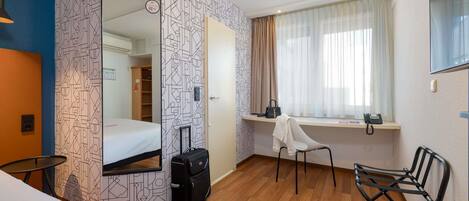 A hotel room with a bed, a desk with a chair, a suitcase, and a window with curtains.