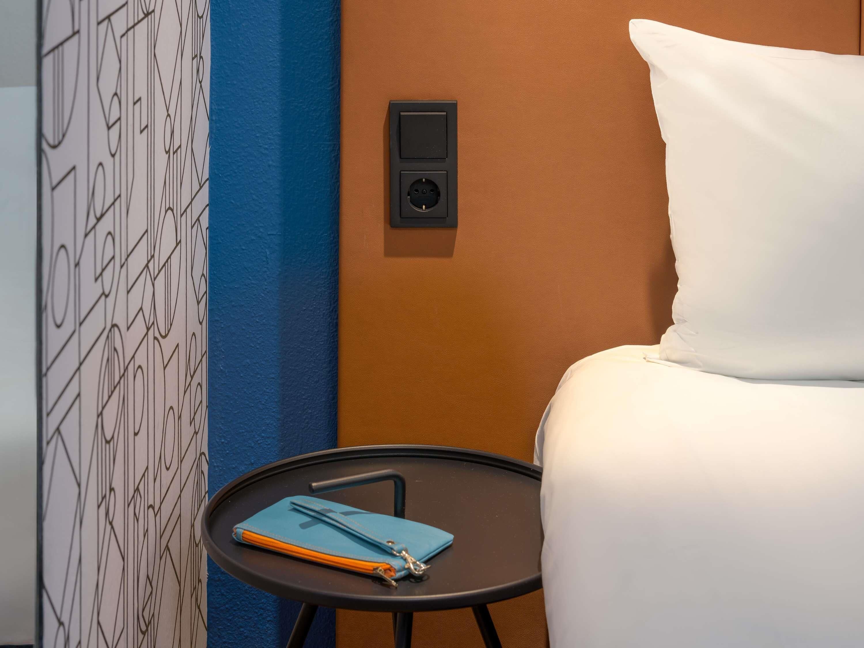 A hotel room with a bed, a bedside table, a power outlet, and a wall with geometric patterns.