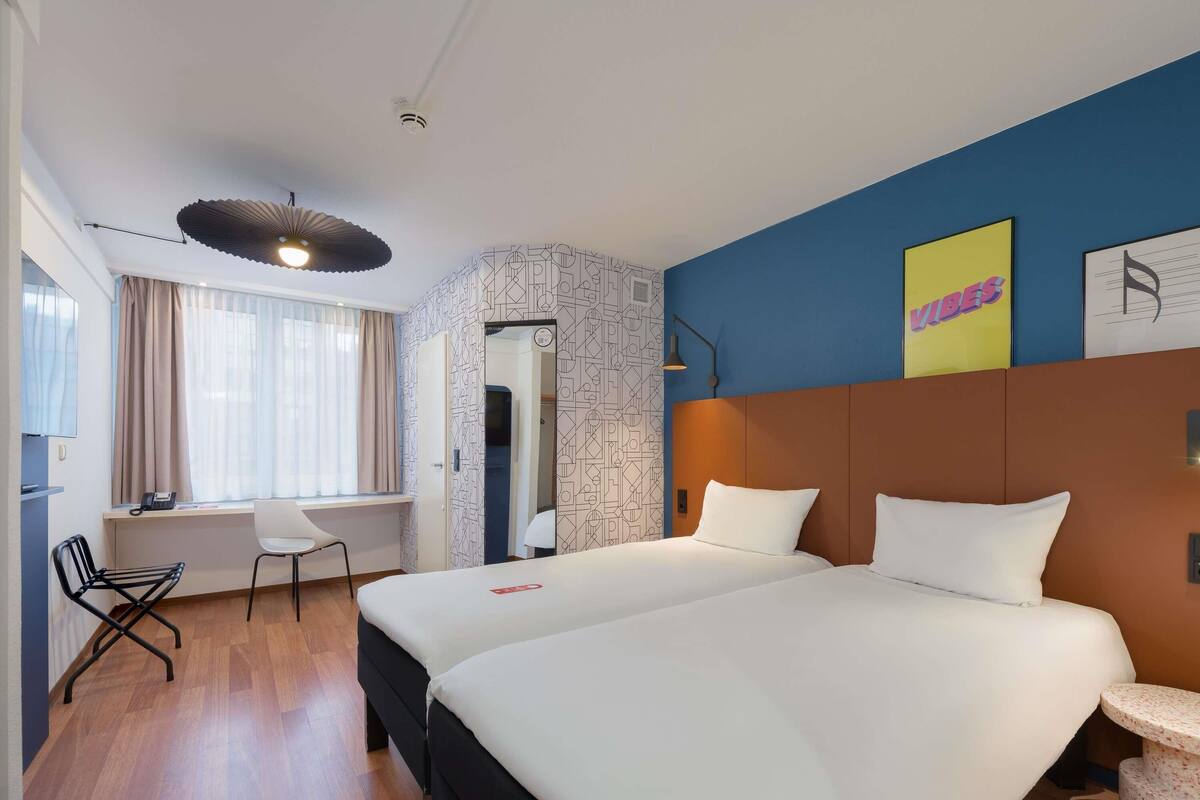 A hotel room with two beds, a desk, a chair, a small table, and a mirror.