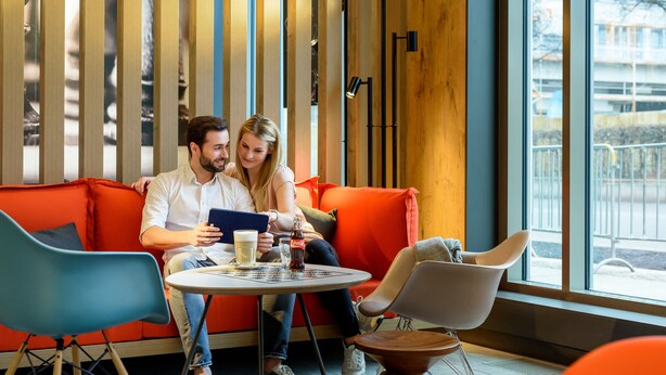 Una pareja sentada en un sofá rojo con una mesa entre ellos, una ventana con vistas y muebles modernos.