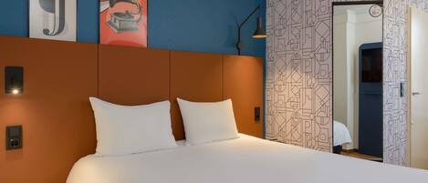Habitación de hotel con cama, mesita de noche, lámpara de pared y puerta con diseño geométrico.