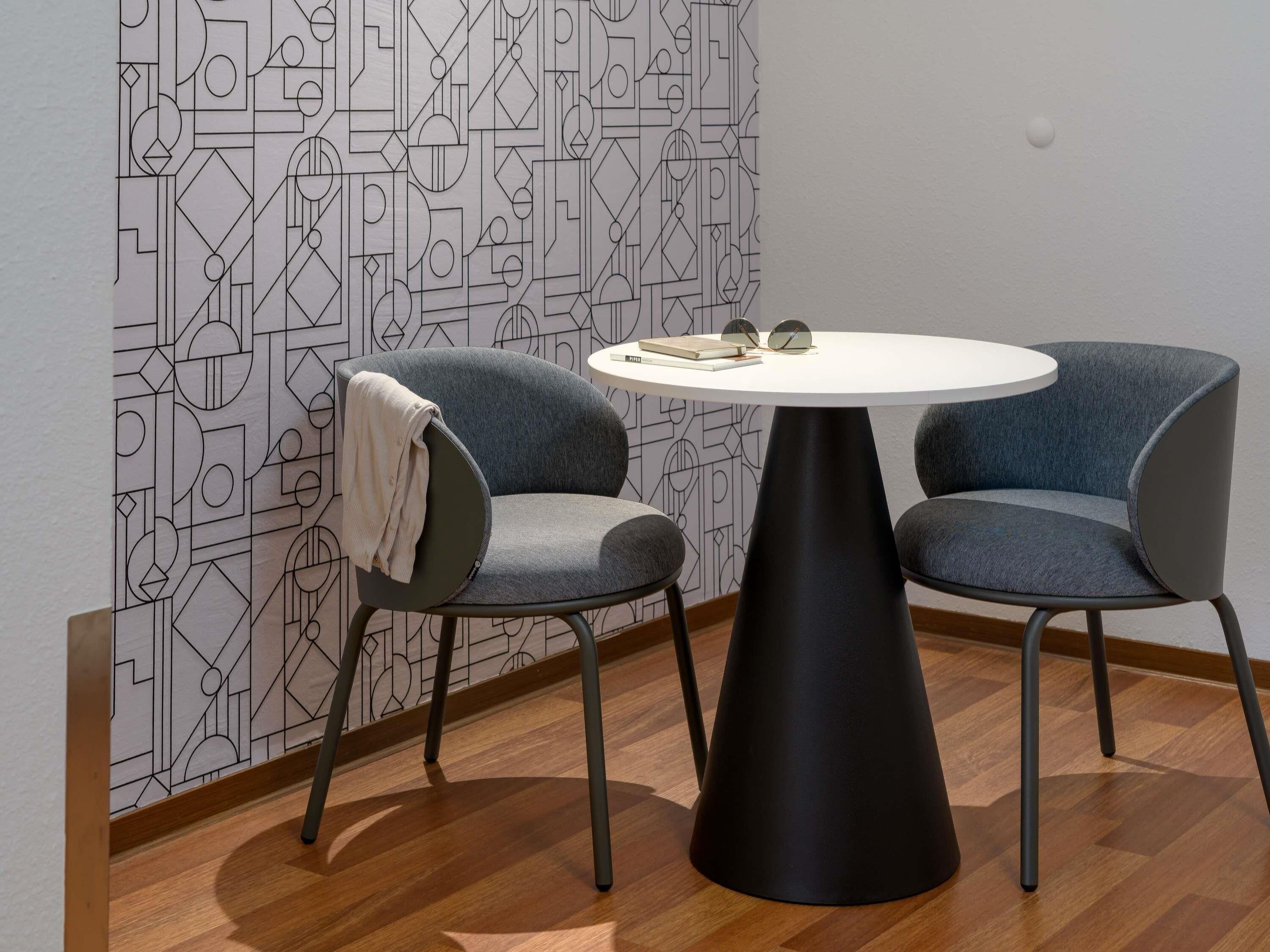 A modern room with a geometric patterned wallpaper, a round table, two grey upholstered chairs, and a black pedestal base.