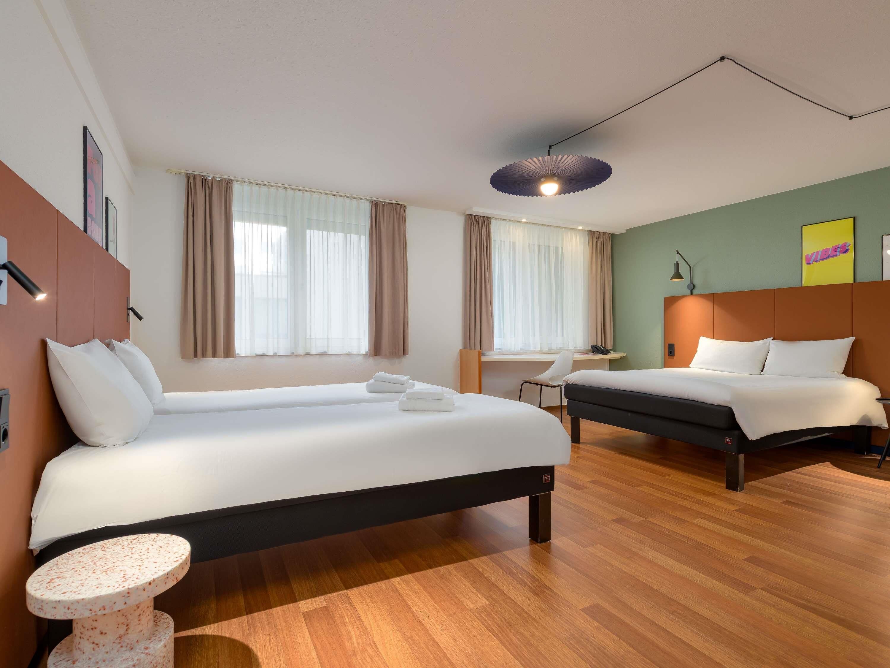 A hotel room with two beds, a desk, a chair, a round table, and a ceiling light.