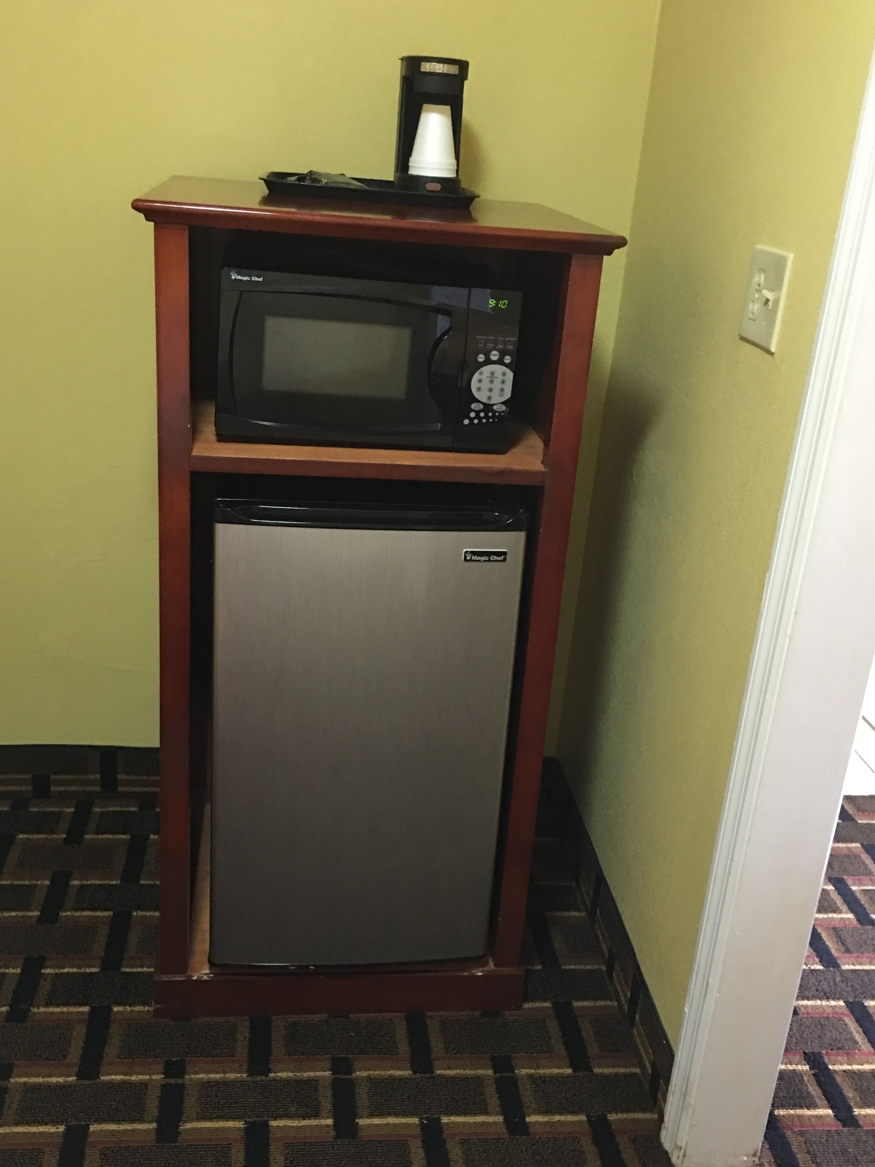 Una pequeña cocina americana con microondas y refrigerador, ubicada sobre un estante de madera frente a una pared amarilla.