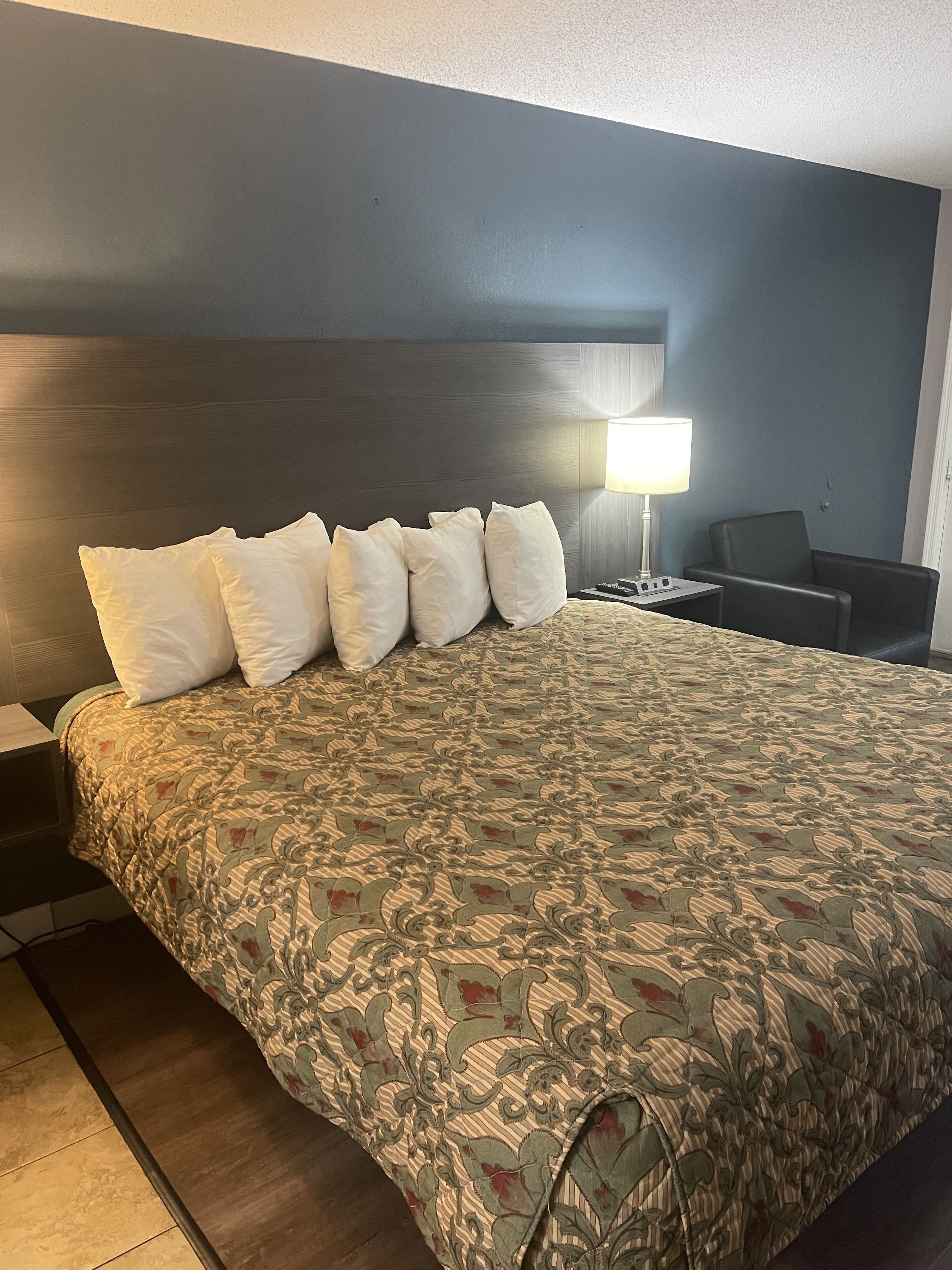 A hotel room with a large bed, a headboard, a bedside table with a lamp, and a chair.