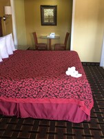 A hotel room with a large bed, a small table with a white coffee maker, two wooden chairs, and a framed picture on the wall.