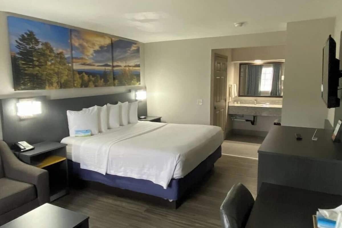 A hotel room with a bed, a desk, a chair, a nightstand, a painting, and a bathroom visible through an open door.