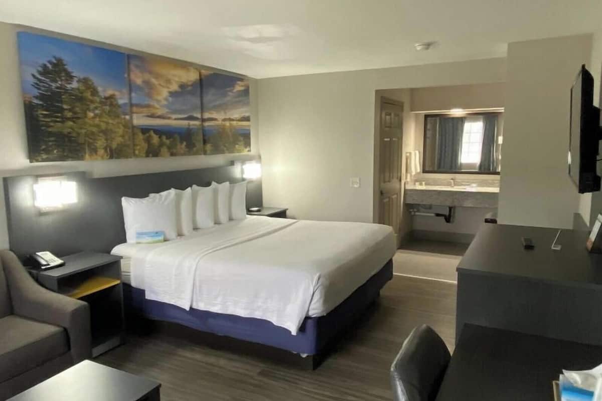 A hotel room with a bed, a desk, a chair, a nightstand, a painting, and a bathroom visible through an open door.