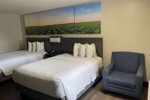 Habitación de hotel con dos camas, un sillón gris y un cuadro grande de un campo en la pared.