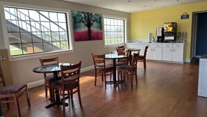 Un comedor con sillas de madera y una mesa de vidrio, ventanas amplias con vistas al exterior, y una zona de cocina con armarios blancos y una cafetera.