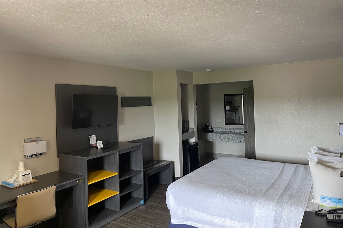 A hotel room with a bed, a desk with a chair, a television, a small shelf, and a bathroom visible through an open door.