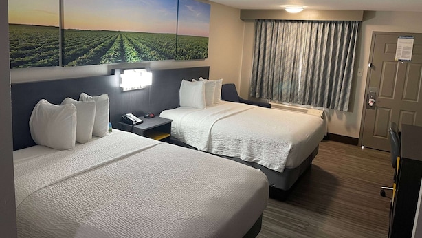 Habitación de hotel con dos camas, un cuadro grande de paisaje en la pared, un escritorio con una lámpara y una puerta con un letrero.