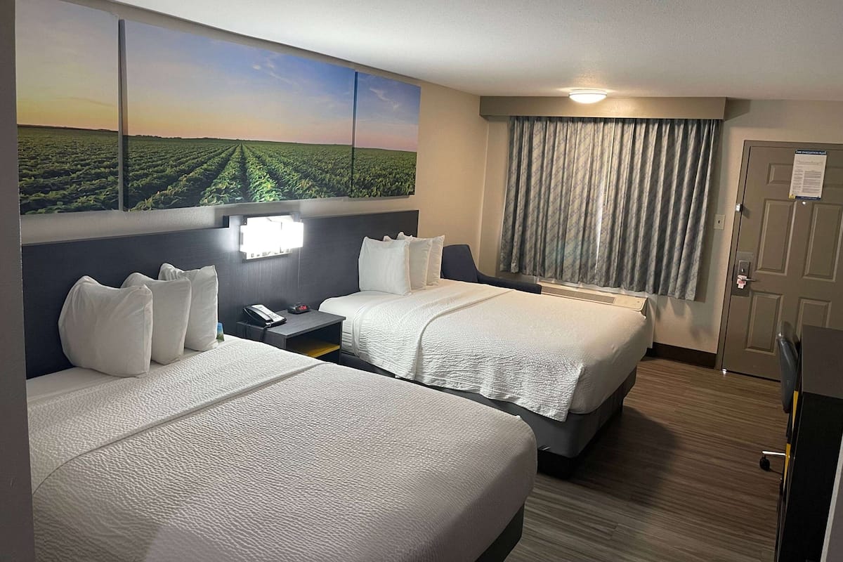 A hotel room with two beds, a large landscape painting on the wall, a desk with a lamp, and a door with a sign.