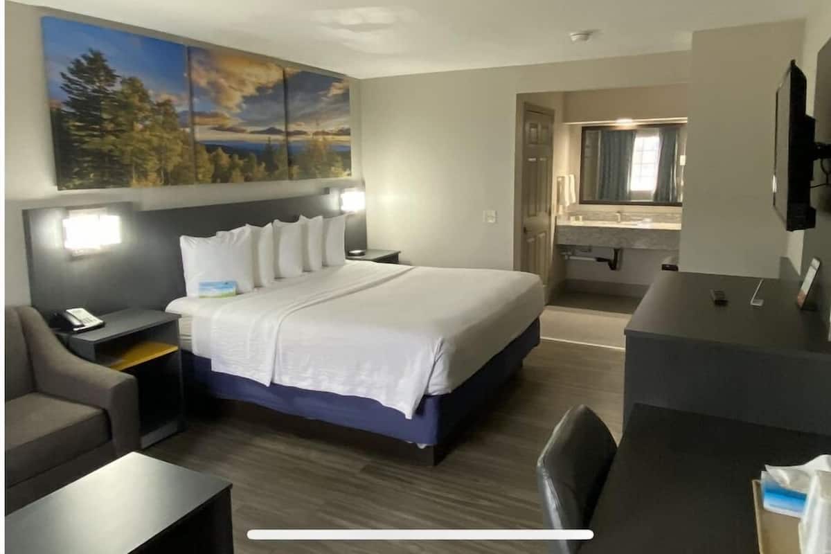 A hotel room with a large bed, a desk, a chair, a nightstand, a painting on the wall, and a bathroom visible through an open door.
