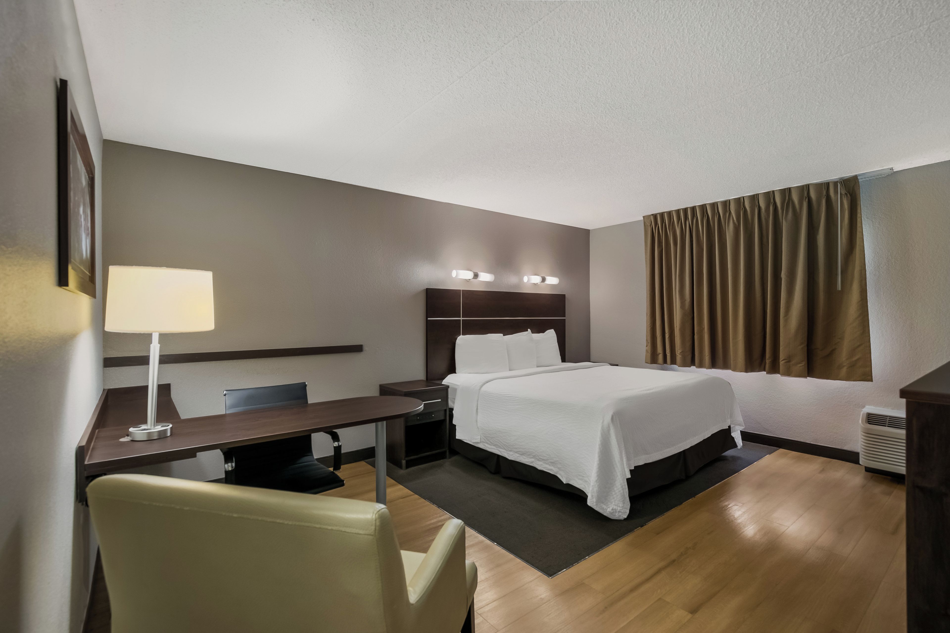 A hotel room with a bed, bedside table, lamp, chair, and a view of the outdoors.