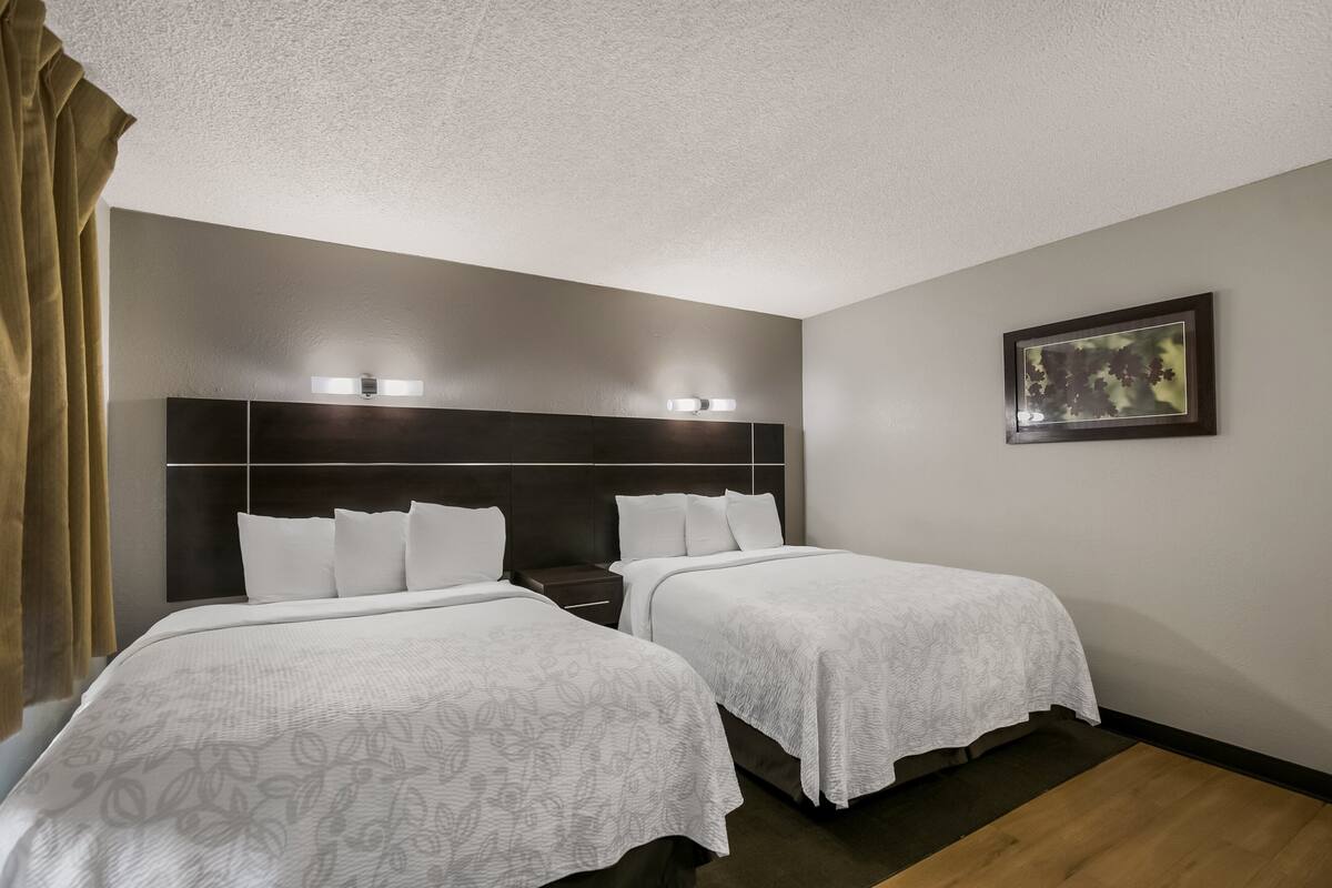 Ein Hotelzimmer mit zwei Betten, einem dunklen Kopfteil, einem gerahmten Bild an der Wand und einem Fenster mit Vorhängen.