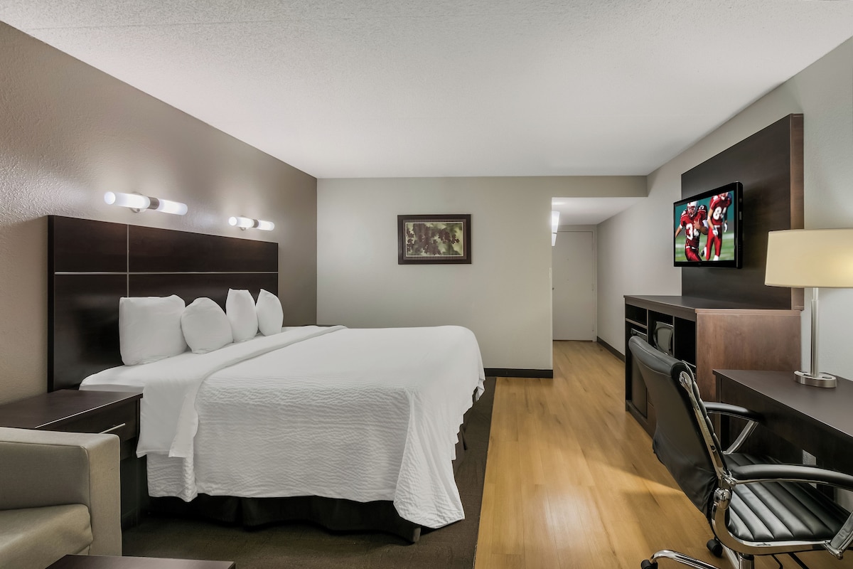 Quarto de hotel com cama, escrivaninha, cadeira, televisão e abajur.